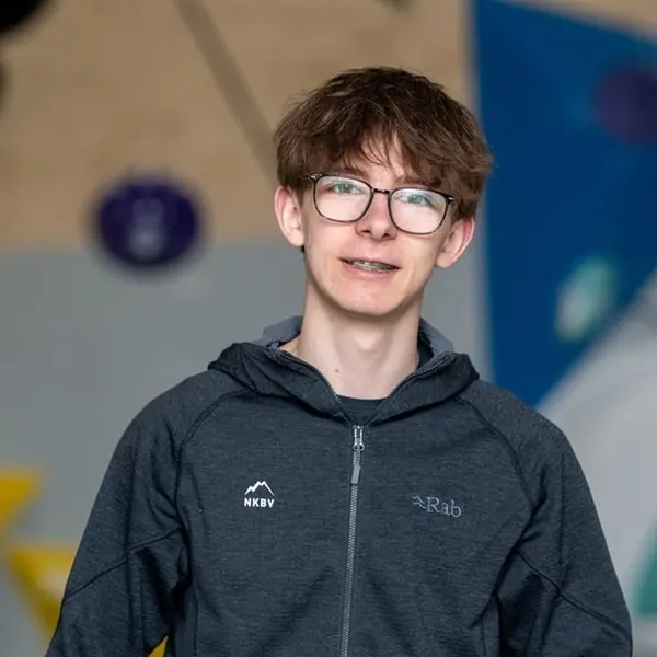 Een jongere met een bril en een beugel, gekleed in een donker jack met ritssluiting, staat binnenshuis tegen een wazige achtergrond met kleurrijke geometrische vormen.